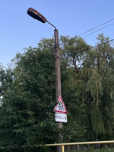 A street lamp with a rusted, elongated lamp head is mounted on a concrete pole in front of lush green trees and a clear blue sky. Below the lamp, a triangular warning sign depicts silhouetted figures running. Underneath this sign is a rectangular sign with an upward-pointing arrow and the text "100 M". The scene is located in Koroviia, Ukraine.