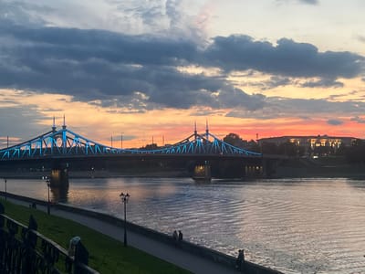 This is a horizontal, eye-level, medium shot of the Starovolzhsky Bridge in Tver, Russia, at sunset. The bridge, illuminated with blue lights, spans a wide river. The sky is a mix of dark, dramatic clouds and vibrant orange and pink hues from the setting sun. The river reflects the colors of the sky and the bridge lights. Along the riverbank, a paved walkway is visible, bordered by a grassy area and a decorative metal fence. A few people are scattered along the walkway, some walking and others sitting by the river. Streetlights line the walkway, and buildings with lights on can be seen on the far bank of the river. The overall atmosphere is serene and picturesque.