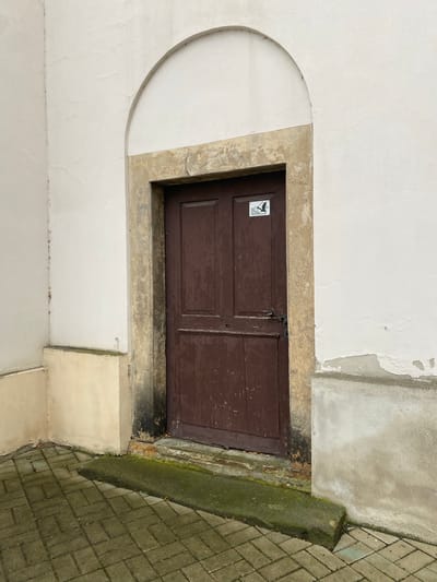 An arched opening frames a weathered, dark brown wooden door set into a cream-colored stucco wall. The door has two panels and a black metal handle. A small white sticker with a black silhouette of a bird in flight and text in an unknown language is affixed to the upper portion of the door. The door is surrounded by a stone archway that is stained and weathered. To the left of the door, the wall transitions to a lighter, creamy yellow color at the base. The ground in front of the door is paved with rectangular grey bricks laid in a herringbone pattern, with a raised concrete threshold covered in moss. The overall scene appears to be an exterior entrance to a building, possibly an older structure, located in Bílina, Czechia. The lighting suggests an overcast day.
