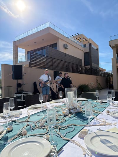 The image shows an outdoor setting in Krimovice, Montenegro, with a band performing in front of a table set for a meal. A modern building with a rooftop terrace and large windows is visible in the background, illuminated by bright sunlight. The table is adorned with a turquoise runner, white plates, champagne flutes, and a nautical-themed centerpiece with seashells and rope. Two men are playing guitars and singing, with musical equipment including speakers and a laptop set up nearby. A third person, partially visible, appears to be part of the band as well. The scene suggests a festive event taking place on a sunny day.