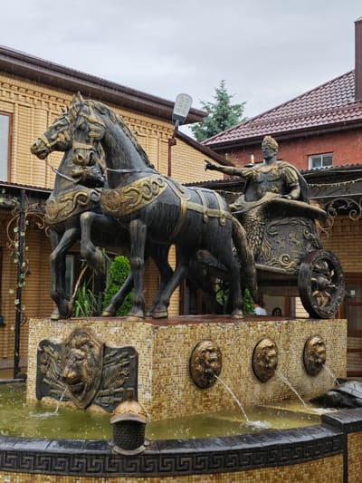 This is a medium shot, outdoor photograph of a bronze sculpture of a Roman chariot being pulled by two horses. The sculpture is part of a fountain and is situated in front of a building with a brick facade. The chariot rider is depicted as a man in Roman armor, pointing forward with his left arm. The base of the fountain features decorative lion heads from which water is spouting, and also a large lion's head relief. The sculpture is dark in color with golden accents on the horses' harnesses and the chariot's details. The weather appears overcast and rainy, with visible raindrops. In the background, parts of a building with a red tiled roof and brown brick walls are visible, along with some greenery. The image is located in Cherkessk, Russia.