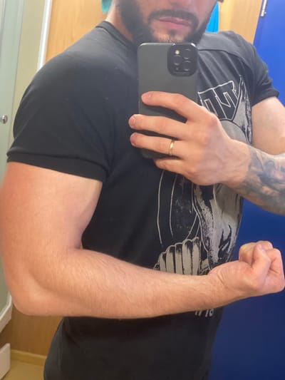 A person is taking a mirror selfie, showing their bicep and forearm. They are wearing a black t-shirt with a graphic print and a gold ring on their left hand. Their right hand is holding a smartphone. The background appears to be a room with a blue door and a light-colored wall. The image is taken in Koskolovo, Russia.