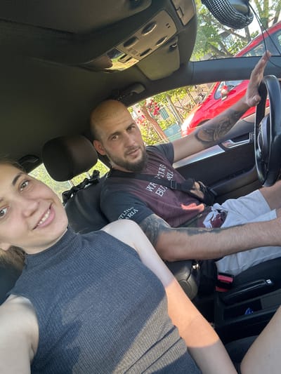 The image captures two individuals, a man and a woman, inside a car cabin, likely posing for a selfie. The perspective is from a low angle, looking upwards at the occupants. The original image is rotated 90 degrees clockwise, so the description assumes an upright orientation.
The man, situated towards the left (driver's side) of the frame, has a short beard and is looking directly at the camera with a calm expression. His arms are visibly tattooed, and he is wearing a dark t-shirt with some white text that partially reads "WE ARE BUILDING BUT..." or similar. A black strap from a bag or accessory crosses his chest. His right arm is extended upwards towards the car's windshield area.
To the man's right, the woman is smiling broadly at the camera. She has dark hair and is wearing a dark, ribbed sleeveless top.
The car's interior is dark-colored, showing parts of the dashboard, a steering wheel, and seat upholstery. Through the windshield, a bright, sunny outdoor scene is visible. A red car is parked directly outside, along with abundant green trees and foliage, and hints of buildings in the background. The bright natural light and visible shadows suggest it is daytime. The scene evokes a relaxed atmosphere, consistent with the specified location context of Rogozen, Bulgaria.