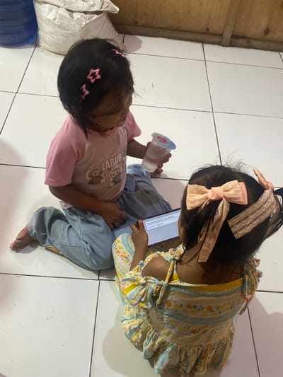 Two young children are sitting on a tiled floor in what appears to be a residential setting in Ta', Indonesia. The child on the left, facing to the right, is wearing a pink t-shirt and light blue jeans. They are holding a clear plastic cup with a blue lid containing a clear liquid. The child on the right, facing away from the viewer and towards the left, is wearing a yellow and white floral-print dress. They are holding a smartphone, looking at the screen. The floor is made of white square tiles. In the background, a large white bag and part of a wooden wall are visible.
