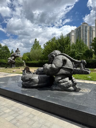 The image shows a bronze sculpture of people on a paved area. The sculpture depicts several figures, including someone carrying a load on their back and another figure holding a circular object. In the background, there are trees, a tall building, and a gazebo-like structure. The sky is partly cloudy. The location is Changping District, China.