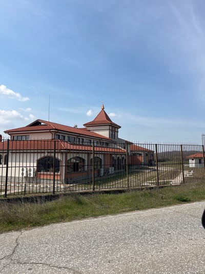 An exterior scene in Zlatovrah, Bulgaria, depicts a multi-story building complex viewed from an adjacent road under a clear blue sky with scattered white clouds. The primary structure features cream-colored walls with a dark brown or red brick base and an extensive, multi-sectioned red-tiled roof. A prominent central tower-like section with a conical red-tiled roof and a spire-like finial is visible. Numerous dark-paned windows, some arched on the ground floor and rectangular on upper levels, are present. Multiple external air conditioning units are affixed to the building's facade. A tall, thin antenna or lightning rod extends from the main roofline.
A black metal fence, constructed from vertical bars, separates the building's property from the foreground, curving towards the right where a dirt or gravel path leads to a smaller, single-story white building with a red-tiled roof, partially obscured by distant trees. The ground immediately behind the fence appears to be a mix of dry earth and sparse grass. The foreground consists of a grey asphalt road surface with visible cracks, bordered by a strip of unkempt green and dry grass and weeds between the road and the fence. No individuals or active subjects are present. The environment appears rural or semi-rural, with trees visible beyond the fenced property to the right.