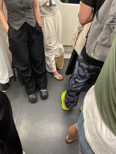 This image shows the legs and feet of several people standing on a gray floor, likely in a public transportation setting like a train or bus in Futian District, China. The perspective is from above, looking down at the individuals' attire and footwear.
On the left, a person wears a grey vest over black pants and black Crocs with white charms. To their right, another person is dressed in a white linen outfit with white sandals. Further to the right, a person sports camouflage-patterned pants and bright lime green Crocs. Another individual, partially visible, is wearing blue denim shorts and brown sandals. The background includes parts of clothing and bags, suggesting a crowded space. The floor appears to be a speckled grey material. No specific time of day or weather conditions are discernible. The overall impression is of people commuting or traveling.