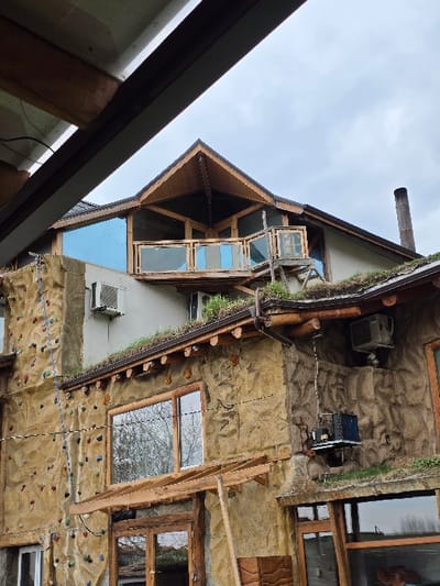 A multi-story building in Slokoshtitsa, Bulgaria, is depicted under an overcast sky, partially framed in the upper left by a dark beam and white gutter from an unseen foreground structure. The building displays an eclectic architectural style. The lower and mid-levels feature a textured, rock-like facade with integrated elements. An outdoor climbing wall, equipped with numerous multi-colored holds, is prominent on the left side of the lower facade. Multiple rectangular windows with wooden frames are visible, reflecting ambient light and distant vegetation. A "green roof," covered with live vegetation, spans the mid-level, bordered by rustic wooden eaves and gutters. Several external air conditioning units are mounted on the building's walls at various heights. The upper section consists of a lighter-colored plastered facade, topped by a dark-shingled pitched roof. An open-air balcony with glass and wood railings is situated on this upper level, partially covered by an angular, open-frame wooden roof structure. A slender, dark metal chimney stack extends upwards from the right side of the main roof. No individuals or explicit actions are present within the frame.