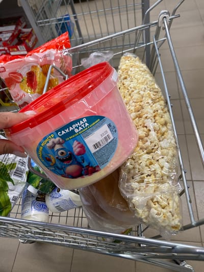 The image shows a shopping cart filled with various grocery items. In the foreground, a person's hand with manicured nails is holding a pink plastic container of cotton candy. The container has a label with blue text and a cartoon monster character. Next to the cotton candy is a large, clear plastic bag filled with popcorn. In the background, other items are visible, including a red bag with strawberries and cake, and several white bottles with blue and white labels. The shopping cart itself is made of metal and is positioned on a tiled floor. The overall scene suggests a person is shopping for snacks. There is no information available about the specific location in Kushnarenkovo, Russia.