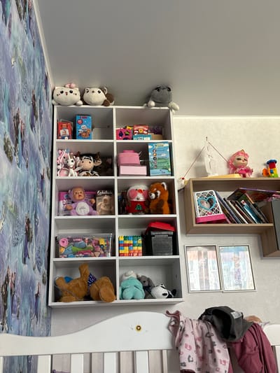 The image displays a white bookshelf filled with children's toys and books, positioned against a wall adorned with a Frozen-themed wallpaper. Two stuffed animal characters are on the top shelf, alongside what appears to be a plush pug. Below them, various toys and boxes are neatly arranged on the subsequent shelves. On the right side of the image, a smaller wooden shelf holds books, a doll with pink hair, and a toy train. Two framed pictures hang on the wall above this shelf. In the foreground, a white crib with bedding and clothing draped over it occupies the lower portion of the frame. The setting appears to be a child's bedroom.