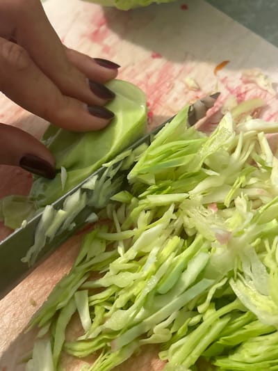 A close-up, overhead shot captures a person's hand with dark red nail polish slicing through a head of cabbage on a wooden cutting board. The knife is mid-cut, creating thin, shredded pieces of cabbage that pile up on the board. The lighting is warm, highlighting the texture of the vegetables and the wood grain. The background is blurred, with hints of pink and white, possibly from other ingredients or the cutting board surface. The scene appears to be in a kitchen, likely for food preparation.