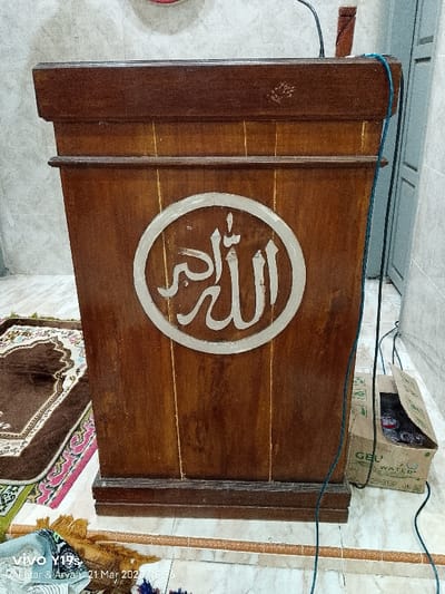 The image shows a wooden podium with a large, white circle in the center depicting Arabic calligraphy. The podium is brown and has a worn surface. The circle has a thin, white rim and the inscription is in a serif font. The podium is located in a room with tiled floors. A prayer rug is visible in the foreground. A cardboard box is lying on the floor next to the podium, containing various objects. The background is partially obscured by a wall. The scene is located in Kecamatan Kroya, Indonesia, at coordinates -6.423 latitude and 107.996 longitude. The image is taken at a slightly low angle, focusing on the podium. The lighting is even and well-lit.