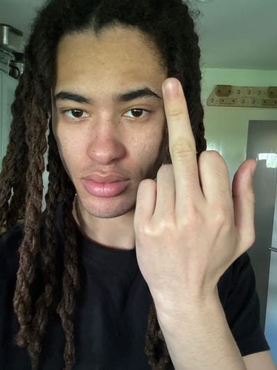 A portrait-style image, rotated 90 degrees counter-clockwise, features a person with dark brown dreadlocks and a medium-dark complexion, looking directly at the camera. The person has dark brown eyes and visible freckles across their face. A faint shadow of stubble or a short beard is present on the chin and upper lip.

The person's left hand is raised in front of their face, making a "middle finger" gesture towards the viewer. The skin tone of the hand and visible arm appears lighter than the face. The individual is wearing a black t-shirt.

The background indicates an indoor setting, likely a room. A light-colored wall, possibly pale green or grey, is visible. In the upper right background, a wooden climbing hangboard or similar training device with multiple holds and drilled holes is mounted on the wall. A window or light source is partially visible, casting some light into the scene.

The lighting suggests an indoor shot, possibly during the daytime. The overall mood is casual and subtly defiant due to the hand gesture, while the facial expression remains relatively neutral. There is no visible text in the image. The context places the location in Les Ulis, France.
