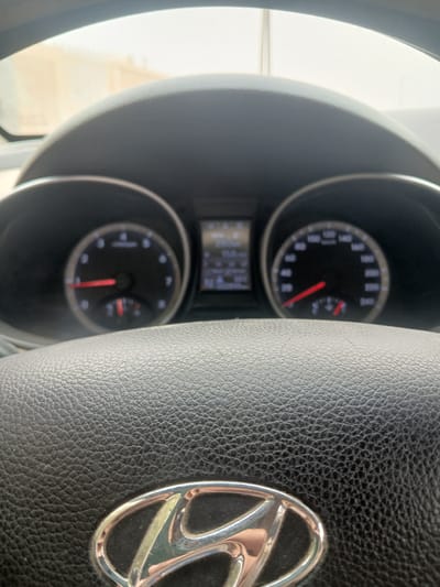 The image shows the interior of a car, specifically the dashboard and steering wheel. In the foreground, a black leather steering wheel with a silver Hyundai logo is visible. Behind the steering wheel, the car's instrument cluster is shown, with a speedometer and tachometer dials. The speedometer needle is pointing towards approximately 60 km/h. The background appears to be the exterior of the car, suggesting it is parked outdoors, with natural light filtering in. The overall setting is consistent with a vehicle parked in an urban or semi-urban environment, such as the city of Hun, Libya. There are no people visible in the image.