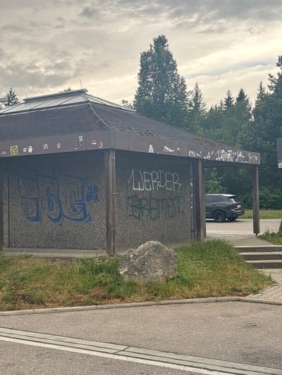 The image shows a covered structure with graffiti on the walls and stickers on the roof overhang. The structure appears to be a bus stop or a small shelter. On the left side of the structure, there is blue graffiti on a textured wall. In the center and right, there is white and green graffiti. The roof is dark and slopes downwards. There are stickers scattered across the roof overhang. A dark-colored SUV is parked to the right of the structure, partially visible through an opening. In the foreground, there is a paved road with white lines. To the left of the structure, there is a large rock and green grass. Behind the structure, there are trees and a cloudy sky. Based on the graffiti "WERDER BREMEN", this location is likely in Heidenheim an der Brenz, Germany, as Werder Bremen is a well-known football club and their name is often used in graffiti in various cities.