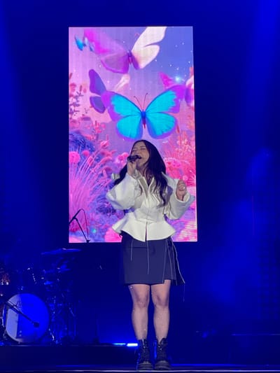 A woman is singing on a stage in front of a large video screen displaying a butterfly image. She is wearing a white top, a dark skirt, and dark boots. She is holding a microphone. To the right of the stage is a drum set. The stage is illuminated with blue lighting. The specific location in Salembaran, Indonesia, cannot be determined from the image.