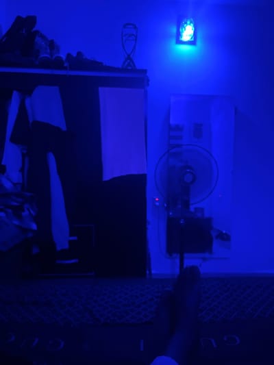 The image depicts a dimly lit room bathed in blue light, with a prominent blue light source on the wall casting a glow. A shelf above holds several pairs of shoes. Below the shelf, clothing items hang from what appears to be a closet or wardrobe. To the right, a fan is visible, and a framed artwork or poster is mounted on the wall. The floor is covered by a patterned rug, and a person's feet are visible in the foreground, resting on the rug. The scene is set in Nabatum, Nigeria.