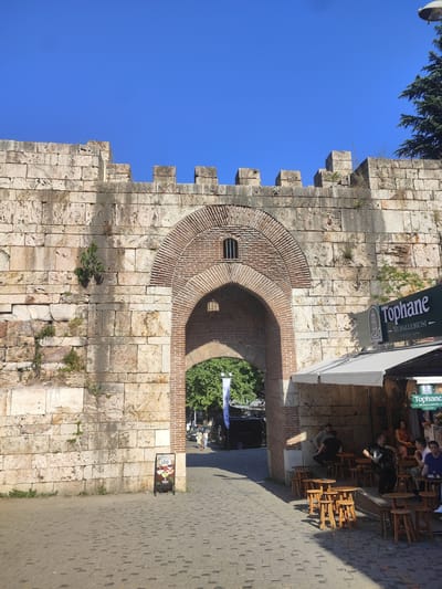The image depicts an arched gateway within a large, weathered stone wall, likely a historical structure. The wall is constructed of rough-hewn, rectangular stone blocks, with some sections exhibiting signs of age and vegetation growth. The archway itself is reinforced with red brickwork in a concentric pattern. Above the arch, a small, barred window is visible within the brick structure. The ground in front of the gateway is paved with cobblestones.

To the right of the archway, a small outdoor seating area is set up with wooden tables and stools. Several people are seated at these tables, appearing to be patrons of a cafe or restaurant. A green sign with white lettering, reading "Tophane Muhallebicisi - Since 1940 -", is prominently displayed above the seating area. Another similar sign is visible further to the right.

In the background, through the archway, trees and a few people walking are visible. A black van is parked just beyond the gateway. The sky above is a clear, bright blue. The overall scene suggests a public space, likely in an urban area, possibly a historical district given the architectural style of the wall and gateway.

Based on the signage and the architectural style, this location is likely Tophane Muhallebicisi, a well-known dessert shop, situated in the vicinity of the historical Tophane neighborhood in Osmangazi, Bursa, Turkey, famous for its Ottoman-era clock tower and structures. The presence of outdoor seating and patrons indicates a lively, functional establishment.