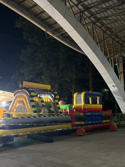 In Koronadal City, Philippines, an outdoor event space is captured at night under a large, modern roof structure. The foreground features a brightly colored inflatable playground, predominantly red, blue, yellow, and green, with black and yellow striped accents on a larger grey inflatable section. Yellow "CAUTION" tape cordons off the inflatable attraction, with several orange anchors visible at its base. Two individuals, one appearing to be an adult wearing a light-colored shirt and hat, and another person partially obscured, are present within the inflatable structure. Behind the playground, trees are visible against the dark night sky. The roof structure above is supported by visible wooden beams and has a white, angular facade.