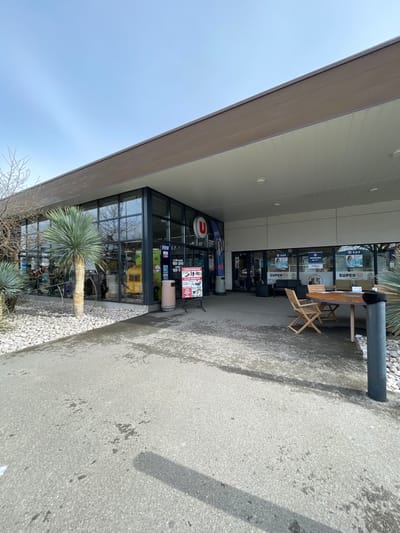 The image depicts the exterior of a Super U supermarket in Puyvert, France, under a clear blue daytime sky. The modern commercial building features a flat, dark brown roofline extending into a white soffit over a sheltered entrance area. The facade is predominantly glass on the left, reflecting bare deciduous trees, while the right portion under the overhang has lighter-colored panels.
In the foreground, an asphalt or concrete paved area shows darker, possibly wet or stained, patches. This pavement gives way to landscaped sections filled with white gravel and several large, spiky-leafed plants.
At the entrance, directly in front of the glass doors, a light-colored cylindrical trash receptacle stands next to a black A-frame sign displaying white text and graphics, advertising "SUPER U" and other items. A tall banner with a woman's image and "SUPER U" branding is also visible. To the right, within the sheltered area, a light-colored wooden table with three matching wooden chairs is arranged on the pavement. A dark, cylindrical bollard-like object is present in the gravel landscaping to the right of the seating area. The interior of the store is partially visible through the glass, showing further advertisements and what appears to be retail shelving or fixtures. No individuals are present or engaged in any activities within the visible scene.