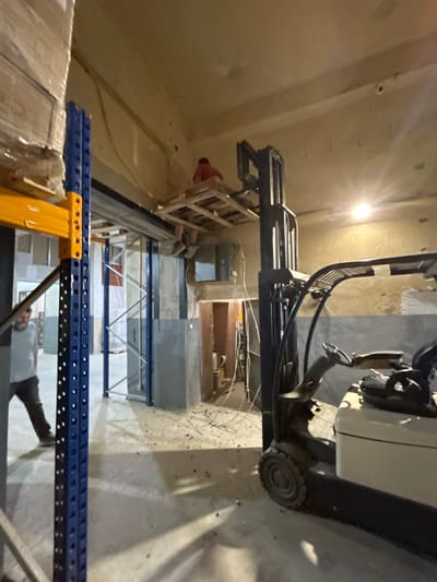 The image depicts an indoor scene, likely a warehouse or industrial space in Djerman, Bulgaria. The primary subjects are a forklift and several storage shelving units. A person is visible operating the forklift, which is positioned with its forks extended upwards, suggesting it is in the process of lifting or moving an object. The shelving units, made of blue and yellow metal, are arranged in rows, indicating a storage area.
The setting appears to be dimly lit, with a bright light source visible in the upper left corner, casting shadows and creating a somewhat stark atmosphere. The walls and ceiling are painted a neutral color, and the floor is concrete. Wires and cables are visible along the ceiling and walls, suggesting ongoing construction or electrical work.
The activity taking place is material handling, with the forklift being used to manage goods within the storage facility. The overall impression is one of work in progress, with the focus on the operational aspects of the warehouse. There is no visible text in the image. The time of day and weather are not discernible from the indoor setting.