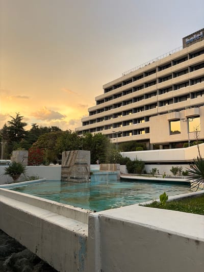 This image captures a modern building and a water feature against a backdrop of a twilight sky. The building, with its repeating rectangular windows and concrete facade, is a prominent subject, suggesting an urban or institutional setting. Next to it, a pool or fountain with clear blue water is visible, decorated with mosaic tile accents. Lush greenery, including trees and smaller plants, surrounds the water feature, adding a natural element to the scene.
The sky is filled with soft, warm hues of orange and yellow, indicating that the photo was taken at dusk or dawn. Wispy clouds are scattered across the sky, catching the fading light. The overall mood is serene and tranquil, with the soft lighting and the presence of water contributing to a peaceful atmosphere.
The location is identified as Sandanski, Bulgaria. There is no visible text in the image. The composition, with the building angled and partially in shadow, and the water feature in the foreground, creates a dynamic and visually interesting perspective. The weathered concrete edges of the water feature suggest it has been in place for some time.