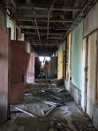 The image shows an interior hallway of an abandoned building, likely in Unknown, Abkhazia. The hallway is in a state of severe disrepair. The ceiling is partially collapsed, with exposed beams and wiring. The walls are peeling and stained, and debris litters the floor. In the center of the hallway, a person wearing a yellow t-shirt and blue shorts stands facing away from the camera, looking down the corridor towards a bright light source. The person appears to be alone. Doors line both sides of the hallway, some open and others closed. The overall atmosphere is one of decay and abandonment.