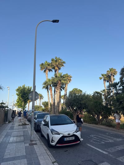In l'Alfàs del Pi, Spain, along a tree-lined street with palm trees, a white car is parked on the right side of the road. The car, a Toyota Yaris, has the license plate "9536 KVF." A woman in a black tank top and denim shorts is standing next to the car, holding a phone to take a photo. Further down the road, a man in a light-colored polo shirt and white shorts is standing on the sidewalk. On the left side of the image, a sidewalk is visible with a gray and white checkered pattern. Several people are walking on the sidewalk, and a line of parked cars is visible behind the white Yaris. The sky is clear and blue, indicating a sunny day. A streetlamp is present on the left side of the image.