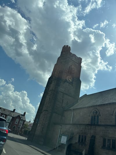 The image shows a low-angle view of Christ Church, Beeston, Nottingham, UK, taken from the street. The church's stone tower dominates the left side of the frame, its clock face visible. Part of the main church building extends to the right. The sky fills the left upper portion of the image, displaying a mix of blue sky and white clouds. Below the church and tower, on the right side of the image, are a glimpse of the street, a red-brick building, and a car.