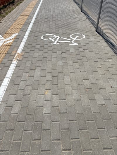The image depicts a paved walkway or bike path constructed of grey interlocking bricks. A white line runs parallel to a strip of textured yellow tactile paving, indicating a designated path for pedestrians or cyclists. A white stencil of a bicycle is painted on the bricks, signifying a bike lane. In the background, a metal chain-link fence runs along the right side of the path. The scene is outdoors, under natural daylight, with patches of dry grass visible to the left of the path. The overall impression is one of urban infrastructure, likely in the city of Smalyavichy, Belarus, given the context. There are no people or animals visible in the image.