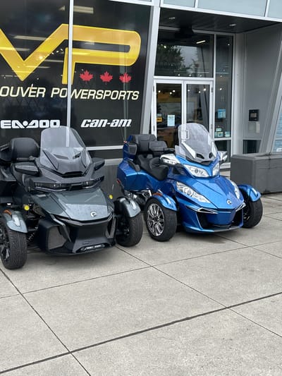 Two Can-Am three-wheeled vehicles, a blue one in the front and a gray one behind it, are parked on a sidewalk in front of a building. The blue Can-Am has a windshield and chrome accents. The gray Can-Am is darker and has a more angular design. Both vehicles appear to be new or well-maintained.
The building behind the vehicles has large windows and a dark facade. A large yellow and black sign on the building reads "VANCOUVER POWERSPORTS" and features three red maple leaves. The "can-am" logo is also visible on the sign.
The sidewalk is made of concrete and is wet, suggesting it has recently rained. The lighting is overcast, indicating a cloudy day. The scene is likely a dealership or a showroom for powersports vehicles. There are no people visible in the image. The time of day appears to be daytime.