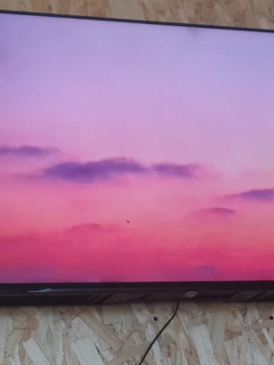 A close-up, slightly blurry, horizontal medium shot captures a television screen displaying a vibrant sunset. The sky transitions from a soft lavender at the top to a deep pink near the bottom. Scattered clouds, colored in shades of purple and pink, are visible in the sky. A tiny, dark, insect-like object is present in the center of the screen, appearing to be flying. The TV screen is mounted on a textured, light-colored surface, possibly oriented plywood, with visible wood grain and knots. The bottom edge of the TV is dark and appears to be a bezel. A black cable extends downwards from the bottom right corner of the TV, suggesting it's plugged into a power source or connected to other equipment, which is not visible in the frame. The scene is devoid of any people or recognizable city landmarks.