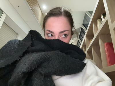 This image, taken in Mundolsheim, France, captures a close-up, low-angle perspective of a young person peeking out from under a dark, textured garment, possibly a sweater or blanket. Only her forehead, eyes, and parts of her cheeks and chin are visible. She has wide, brown eyes and is looking upwards and to her left (the viewer's right if the image were correctly oriented), conveying a curious, playful, or surprised expression. She wears gold hoop earrings.
The background reveals an indoor setting. To the left of the subject, a light-colored wooden bookshelf or shelving unit is visible, partially filled with books, one of which is a prominent red-orange volume. Above the subject's head, a white, possibly fuzzy, garment or blanket is draped. To the right, a window or glass panel with horizontal blinds is visible, set within clean white walls, suggesting a modern interior. A subtle light fixture can be seen in the lower left corner. The lighting indicates it could be daytime or early evening. No legible text is visible within the frame.