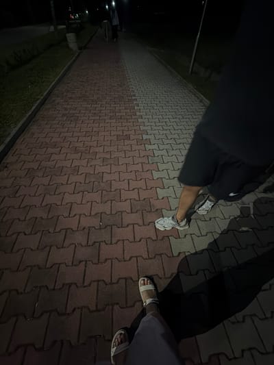 The image depicts a nighttime scene on a paved walkway, likely in the city of Pravets, Bulgaria, given the provided context. The walkway is constructed of interlocking reddish-brown and grey pavers. To the left of the walkway is a grassy area, bordered by a concrete curb. In the background, a light pole is visible, and further in the distance, other figures and faint lights suggest the presence of a park or public area.

In the foreground, the feet and lower legs of two individuals are visible. One person is wearing white sandals and light-colored trousers. The other individual is wearing athletic shoes and dark shorts. The person in athletic shoes is partially obscured by shadow cast by what appears to be a person standing out of frame to the right. The image is characterized by low light conditions, with shadows playing a significant role in defining the scene. The overall impression is of people walking outdoors at night.