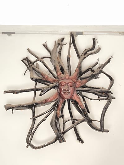 The image shows a handcrafted wall decoration made of wood, resembling a sun with a human face at its center. The face is carved with closed eyes, a nose, and a mouth, and it is surrounded by a circular arrangement of gnarled branches that extend outwards like sun rays. The branches are painted dark brown and black, with some reddish-brown accents on the parts closest to the face, which also appears to be made of wood and painted in a similar reddish-brown hue. The decoration is mounted on a plain white wall, and a faint horizontal pipe or conduit is visible above it. The overall aesthetic is rustic and somewhat primitive. This object is not located in Goron Talo, Indonesia, as no information about its location is available.