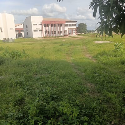 The image depicts a wide view of an academic facility, likely a university campus, in Wukari, Nigeria. In the background, a white building with a red trim is visible, bearing the inscription "FACULTY OF BIOSCIENCES". Several smaller white buildings are also present. The foreground is dominated by a large, overgrown grassy field with a dirt path winding through it. A large tree with dark green leaves is visible on the right side of the frame, partially obscuring the sky. The sky itself is cloudy. The overall scene suggests an outdoor environment on a partially overcast day.