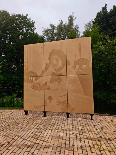 The image displays a large, square sculpture made of perforated panels, arranged in a 3x3 grid. The sculpture is light brown in color and depicts a black and white portrait of a woman, rendered through the perforations. The woman is looking directly at the viewer and appears to be wearing glasses and a bow tie. The sculpture is supported by four dark-colored legs. It is situated outdoors on a paved area made of light brown bricks, with some small green plants growing between the bricks. Behind the sculpture, a dense line of green trees and foliage is visible against an overcast sky. To the left, a black metal fence with decorative finials is partially obscured by the trees and grass. This sculpture is located in the city of Khotkovo, Russia, specifically in the memorial complex of the Radonezh Settlement, dedicated to the writer and ethnographer Lyudmila (Lyubov) Yakovlevna Serebryakova.