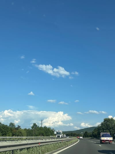 The image shows a view from a vehicle traveling on a highway in Lozen, Bulgaria. The sky is a clear, bright blue with scattered white clouds. On the right side of the highway, there is a dense line of green trees, and beyond that, a rolling hill covered in vegetation. The highway itself has multiple lanes and is bordered by a metal guardrail and a grassy shoulder with some red markers. Several vehicles are visible on the road, including white trucks and cars. One truck in the foreground appears to be a white semi-truck, and another in the distance is pulling a trailer with a blue tarp. The sun is high in the sky, suggesting it is daytime, likely midday given the bright lighting and clear conditions. There is no visible text in the image. The overall impression is one of travel on a sunny day through a scenic, rural landscape.