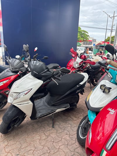 The image shows several parked scooters on a paved surface, likely a sidewalk, in Playa del Carmen, Mexico. A deep blue wall is visible on the left side of the image. Several scooters are grouped together, overlapping each other, obscuring the full view of individual vehicles. The scooters are of varying colors and designs, including red, white, black, and turquoise. One scooter visible in the top right is red and white with the branding "Vitalia 150". Another scooter has "Honda" branding. The scooters appear to be parked near an urban commercial area, with building facades and power lines visible in the background. The sky is overcast.