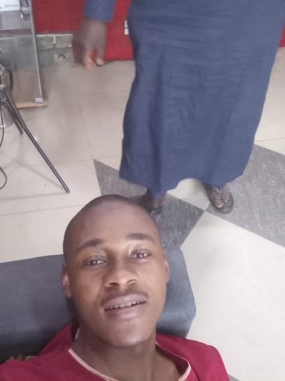 The image depicts an indoor scene in Dorayi, Nigeria. In the foreground, a male subject, appearing to be a young adult, is shown from the chest up, positioned at a low angle. He has a shaved head, light mustache, and is wearing a red garment with a light-colored trim around the neck. His mouth is slightly open, revealing teeth, and he is looking directly at the camera. He is seated on or leaning against a dark gray upholstered surface.
In the background, a second individual stands upright, largely obscured, with their lower torso, legs, and left arm visible. This individual is wearing a long, dark blue dress or robe that extends to their ankles and dark sandals. The floor is tiled with a geometric pattern of light beige and dark gray or black sections. A red wall or partition is visible in the upper right background. On the far left, a metallic cabinet or appliance and a black tripod stand with a visible cable are partially present. No direct interaction between the two individuals is discernable.
