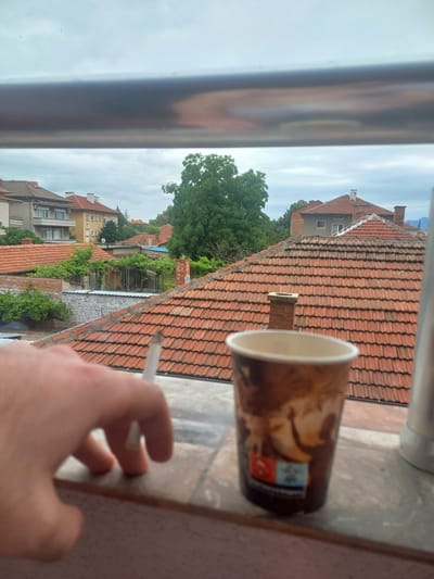 In an outdoor urban setting, a close-up shot frames a hand holding a lit cigarette. To the right, a disposable coffee cup with a busy pattern is positioned on a windowsill. The background reveals a panorama of red-tiled rooftops and buildings under an overcast sky, suggesting a residential area in Parvomay, Bulgaria. The overall atmosphere is casual and contemplative.