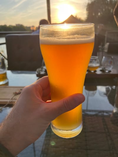 A close-up, first-person perspective shot captures a hand holding a tall glass of golden beer with a foamy head. The beer is condensation-covered, indicating it is cold. In the background, the setting sun casts a warm glow over a blurred landscape of trees and buildings, with the sun's rays reflecting off a body of water. The scene suggests an outdoor setting, possibly a restaurant or cafe with a view. The location is identified as the city of Azov, Russia.