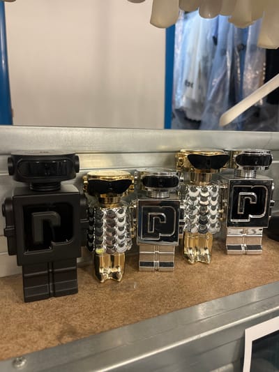 The image displays a row of five elaborately designed containers, likely perfume bottles or similar luxury product packaging, arranged horizontally on a light brown, possibly wooden or cardboard, surface. The image itself appears rotated 90 degrees clockwise, so the objects, which are presumably standing upright, are seen lying on their sides.

From the bottom to the top of the image (which would be from left to right if the image were rotated counter-clockwise to the correct upright orientation):

1.  **Bottom-most object:** This container is entirely matte black. It features a prominent, stylized letter "G" in black on its front face. The body is squarish or rectangular, and it has a matching black cap (appearing on the left side in the current orientation) and a segmented black base (on the right side).
2.  **Second object from the bottom:** This container has a shiny silver-colored body with a distinctive, scaly or textured pattern covering most of its surface. Its cap (left side) and base (right side) are a glossy gold color.
3.  **Third object from the bottom (center):** This container features a glossy chrome or silver-colored body. It displays the same stylized "G" symbol as the all-black object, rendered in black against the silver background. It has a black cap (left) and a silver-colored, segmented base (right).
4.  **Fourth object from the bottom:** Similar to the second object, this container also has a textured, scaly silver-colored body with a glossy gold cap (left) and base (right).
5.  **Top-most object:** Similar to the third object, this container has a glossy chrome or silver-colored body with a black stylized "G", a black cap (left), and a silver-colored, segmented base (right).

The objects are positioned on a flat, light brown surface that shows some texture, possibly a workbench or shelf. A metal channel or rail, likely part of a shelving unit, runs along the left edge of this surface.

In the background, to the far left of the image (top-left if rotated correctly), out-of-focus elements include several white, irregularly shaped objects, which could be plastic components or molds. Below these, there are items wrapped in clear plastic, possibly products or materials, resting on a blue surface. A portion of a white, curved object, possibly a handle or a hook, is also visible extending from the very top-left of the frame.

The overall scene suggests an industrial, manufacturing, or storage environment, likely a quality control station or a staging area for product assembly/packaging. The lighting appears artificial, creating reflections on the highly polished metallic surfaces of the containers. No specific time of day or discernible text beyond the stylized "G" is visible.
