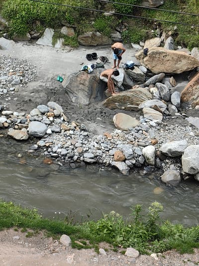 Two young men are visible in the image, on a rocky, muddy riverbank near Kothi, India. One man, wearing blue and white checkered shorts, is bent over, appearing to adjust his clothing. Another man, in light-colored shorts, is crouched near the water's edge, with his hands in the mud. A pile of clothes, including a green and white garment, is situated on a large rock. Several pairs of footwear, including black sandals and green flip-flops, are scattered on the ground. The river flows in the foreground, with grassy vegetation and rocks along its banks. The environment is natural and rugged, with a dominant presence of stones and earthy terrain.