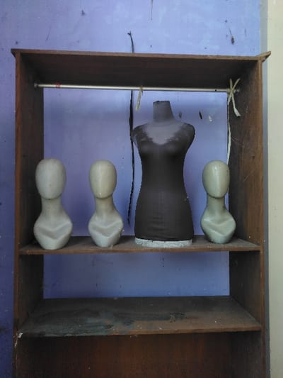 The image depicts a multi-shelf wooden cabinet containing four mannequins, positioned against a faded light purple wall in Elverum, Norway. The cabinet features three visible shelves and a silver-colored metal hanging rod in its upper section. On the middle shelf, one dark gray fabric-covered female torso dress form stands centrally, flanked by three smaller, light-colored, unfeatured neck-and-bust mannequins. The wooden cabinet exhibits signs of age and wear, including dust accumulation on its surfaces. The background wall shows discolored patches and dark smudges, with thin strings or wires hanging in the upper frame and one light-colored string tied to the right end of the hanging rod. No people, actions, or interactions are visible.
