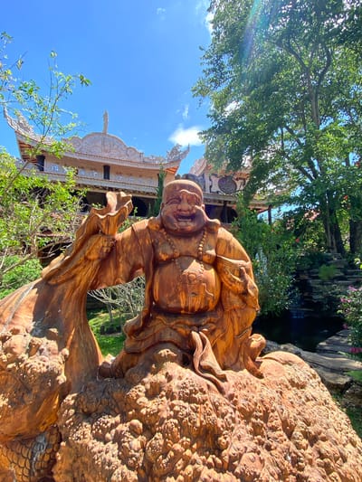 This media file captures a close-up view of a large, intricately carved wooden sculpture of a jovial, perhaps Buddhist, figure. The sculpture is rendered in warm, reddish-brown tones, with detailed textures suggesting flowing robes and beaded necklaces. The figure's face is round and smiling, with closed eyes, conveying a sense of peace and contentment. The sculpture is positioned outdoors, with lush green foliage and trees forming a backdrop.
Behind the sculpture, a portion of a building with ornate, traditional architecture is visible. The building features multiple tiered roofs with decorative trim and what appear to be carved motifs. The overall scene is bathed in bright sunlight, suggesting it is daytime. The sky is a clear, vibrant blue with scattered white clouds, indicating pleasant weather.
The lower right of the image focuses on a textured section of the sculpture that resembles scales or a rough, organic surface, possibly part of the larger structure the figure is integrated with. The wooden carving appears to be a significant artistic or religious artifact, possibly situated within a temple, garden, or other culturally significant site. The image's orientation is vertical, suggesting it was taken with the camera held in portrait mode. The scene evokes a sense of serenity and appreciation for craftsmanship. There is no visible text in the image. The location context provided is Thôn 5, Vietnam, which aligns with the architectural style and potential cultural subject matter.