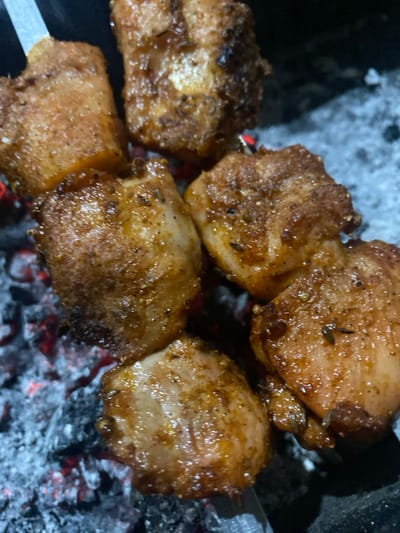 The media file captures a close-up of skewered meat grilling over hot coals, likely in the evening or night due to the prominent glow of the embers and dim ambient light. The scene is set in Abad Tukimachi, Uzbekistan, suggesting a traditional grilling method.
The primary subjects are several pieces of seasoned meat, possibly chicken or lamb, threaded onto metallic skewers. The meat appears golden-brown with some areas of darker charring, indicating it is actively cooking. Visible spices or herbs are sprinkled on the surface of the meat, contributing to its textured appearance. Below and around the meat are glowing red and grey-white coals, some actively burning with a reddish-orange hue, while others are covered in ash.
The activity taking place is outdoor grilling or barbecuing, specifically cooking shish kebabs over an open charcoal fire. The perspective is very close to the grill, emphasizing the sizzling meat and the intense heat from the coals. No people are visible in the frame, but their presence is implied by the preparation of the food.
Notable details include the warm, inviting glow emanating from the coals, which illuminates the underside of the meat. The skewers are partially visible on the left and right edges of the frame, holding the meat pieces together. The lack of bright daylight further suggests a nighttime or late evening setting, where the coals' luminescence becomes a central visual element. There is no visible text in the image.