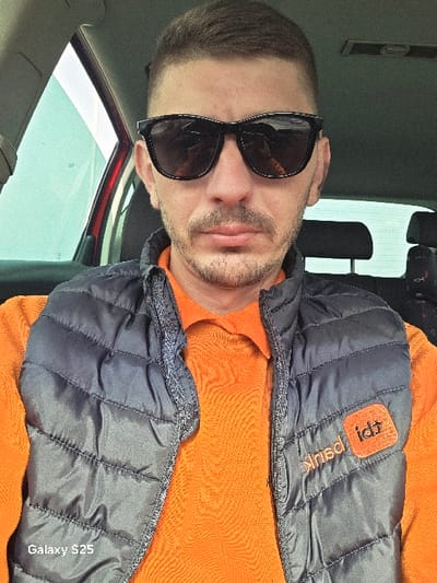 An adult male is depicted in a close-up, front-facing portrait, seated within a vehicle. The location is specified as Novo Delchevo, Bulgaria. The subject has short, dark hair, a trimmed beard, and a mustache. He wears dark, rectangular sunglasses. His attire consists of a vibrant orange collared shirt and a dark grey quilted puffer vest. An orange patch on the subject's right side of the vest displays white text, which includes "idt" and a seemingly mirrored word such as "kirnd". The interior of the vehicle is partially visible, showing a red door frame on the left, a dark-colored seat headrest behind the subject, and a light-colored ceiling. Distant, bright, and indistinct outdoor views are visible through the vehicle's windows. The image features a "Galaxy S25" watermark in the lower-left corner.