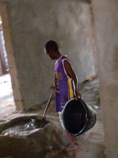 An individual is depicted within an unfinished interior space or construction site in Enugwu-Ukwu, Nigeria. The subject, appearing male, wears a purple and yellow/gold sleeveless basketball jersey with "LAKERS" and the number "36" visible on the front. The person is bent over, actively stirring a dark, liquid mixture on the dirt ground with a long-handled tool, resembling a shovel or hoe. Concurrently, the subject holds an empty, dark-colored plastic bucket in their right hand. The environment consists of rough, unpainted light gray walls and an uneven dirt floor. A vertical structural element, possibly a column, is visible on the left side of the frame. The lighting is dim, suggesting an internal area with limited illumination.
