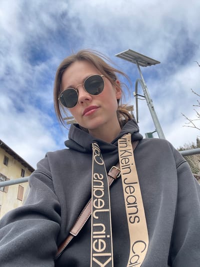 A young woman poses for a selfie in Sappada, Italy. She wears round, dark sunglasses and a gray hoodie with Calvin Klein Jeans straps. In the background, there's a street lamp against a blue sky with clouds and a building.